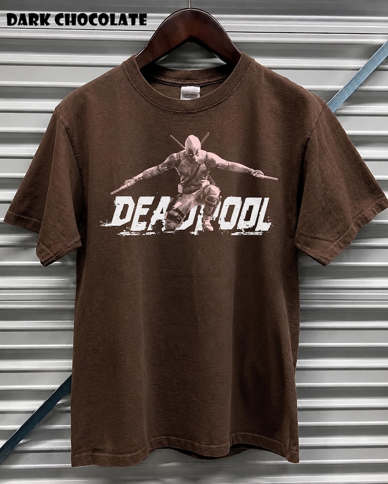 May include: Dark chocolate brown t-shirt with a Deadpool graphic and the word "DEADPOOL" in white. The t-shirt is displayed on a wooden hanger against a corrugated metal background.