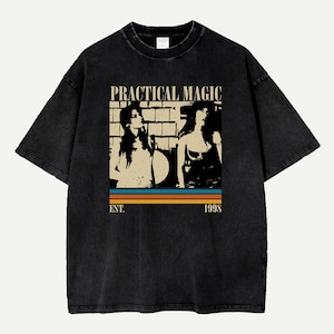Puede incluir: Camiseta negra con un diseño gráfico. El diseño incluye el texto "PRACTICAL MAGIC" sobre una imagen en tonos sepia de dos mujeres. Debajo de la imagen hay tres rayas horizontales y el texto "EST. 1998". La camiseta tiene mangas cortas y cuello redondo.