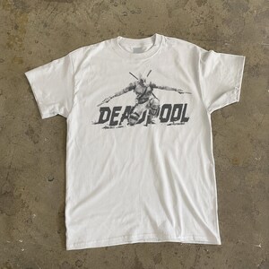 May include: White t-shirt with a grey graphic of Deadpool kneeling, arms extended, holding swords. The word "DEADPOOL" is printed in a distressed font below the character.