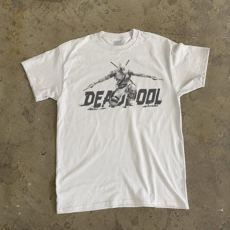May include: White t-shirt with a grey graphic of Deadpool kneeling, arms extended, holding swords. The word "DEADPOOL" is printed in a distressed font below the character.