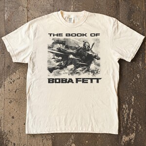 May include: Cream-colored t-shirt featuring a black and white graphic of Boba Fett from "The Book of Boba Fett." The text "THE BOOK OF" is above the image, and "BOBA FETT" is below. The shirt has a crew neck and short sleeves.