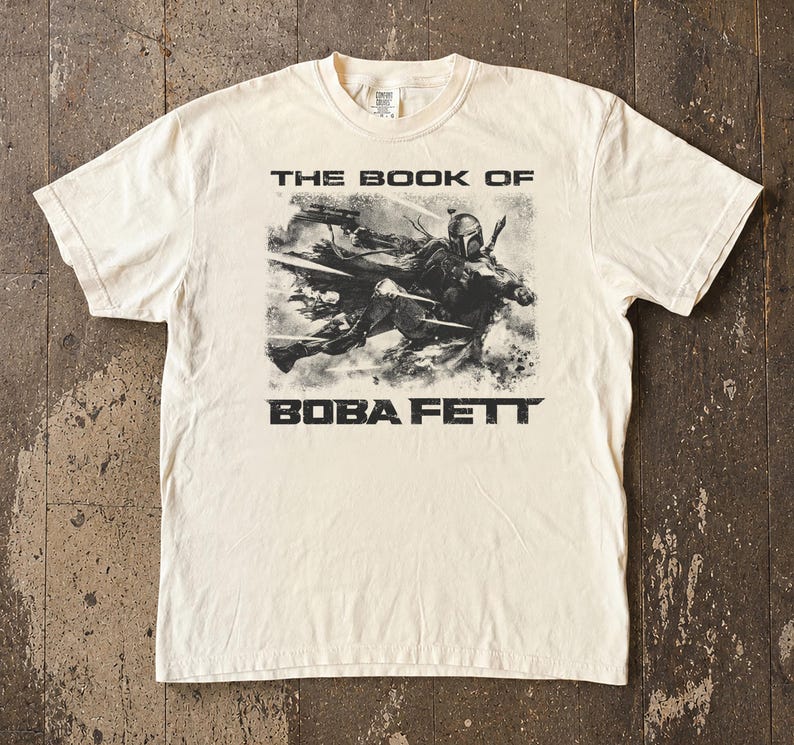 May include: Cream-colored t-shirt featuring a black and white graphic of Boba Fett from "The Book of Boba Fett." The text "THE BOOK OF" is above the image, and "BOBA FETT" is below. The shirt has a crew neck and short sleeves.