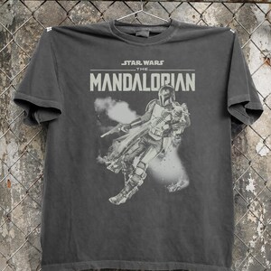 May include: Dark grey t-shirt with the Star Wars: The Mandalorian logo and a graphic of the Mandalorian character. The design is in grey and white, with a distressed, vintage appearance.