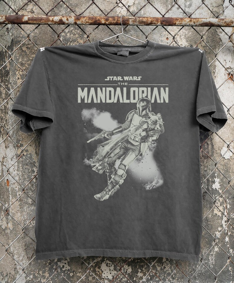 May include: Dark grey t-shirt with the Star Wars: The Mandalorian logo and a graphic of the Mandalorian character. The design is in grey and white, with a distressed, vintage appearance.