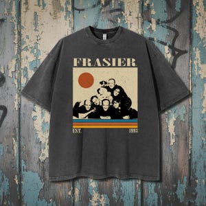 May include: Dark gray t-shirt featuring the word "FRASIER" above a graphic of the cast with a sun and stripes. The text "EST. 1993" is below the graphic. The shirt has a vintage, worn look.