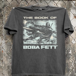 May include: Dark gray t-shirt featuring a graphic from "The Book of Boba Fett." The design shows Boba Fett in action with the text "THE BOOK OF" above and "BOBA FETT" below the image in a light blue color.
