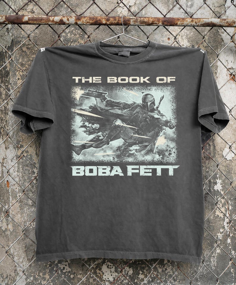 May include: Dark gray t-shirt featuring a graphic from "The Book of Boba Fett." The design shows Boba Fett in action with the text "THE BOOK OF" above and "BOBA FETT" below the image in a light blue color.