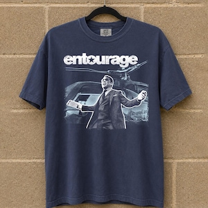 May include: Navy blue t-shirt featuring the word "entourage" in white block letters above a black and white image of a man in a suit with a helicopter in the background. The shirt is hanging on a black hanger.