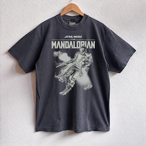 May include: Dark grey t-shirt with a Star Wars The Mandalorian graphic. The design features the text "STAR WARS THE MANDALORIAN" in white, and an image of the character. The t-shirt is displayed on a wooden hanger.