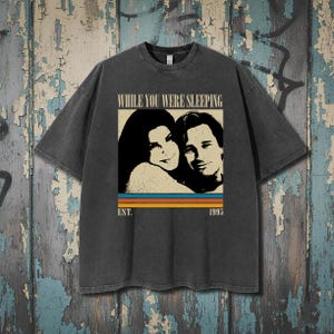 Puede incluir: Camiseta gris oscuro con un gráfico de la película "Mientras dormías". El diseño incluye una imagen en blanco y negro de dos personas, con el texto "WHILE YOU WERE SLEEPING" encima de la imagen y "EST. 1995" debajo.