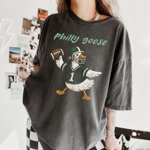 Puede incluir: Una camiseta de manga corta gris carbón con un gráfico de un ganso blanco que lleva una camiseta de fútbol verde y blanca con el número 1 y un casco, sosteniendo un balón. El texto Philly goose está impreso en verde claro sobre el ganso.