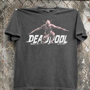 May include: Dark grey t-shirt with a graphic of Deadpool in a crouching pose with two swords. The word "DEADPOOL" is printed in white, distressed lettering across the front. The t-shirt is hanging on a metal hanger.