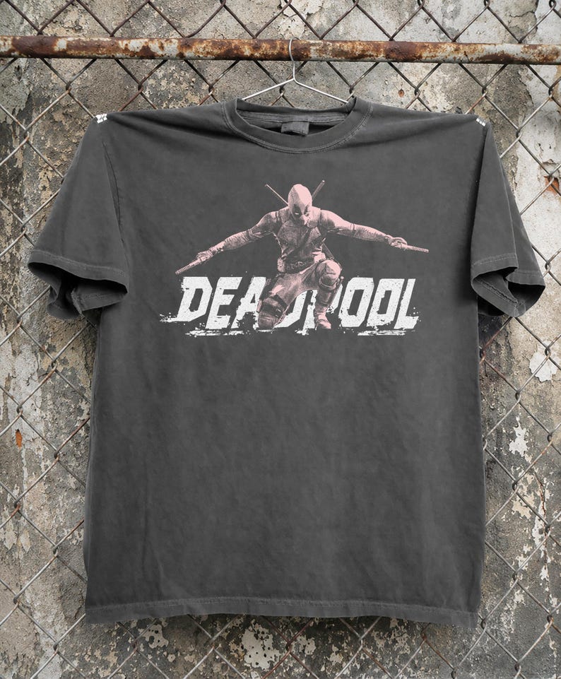May include: Dark grey t-shirt with a graphic of Deadpool in a crouching pose with two swords. The word "DEADPOOL" is printed in white, distressed lettering across the front. The t-shirt is hanging on a metal hanger.