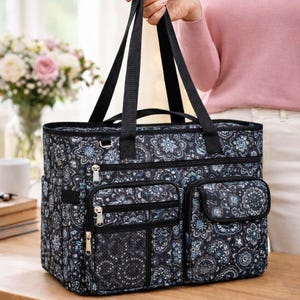 Op de afbeelding: Een zwarte draagtas met een blauw en wit bloemen-paisleypatroon. De tas is voorzien van twee zwarte schouderbanden, een groot hoofdvak met ritssluiting, twee voorzakken met ritssluiting, een voorvak met klep en een zijvak van gaas.