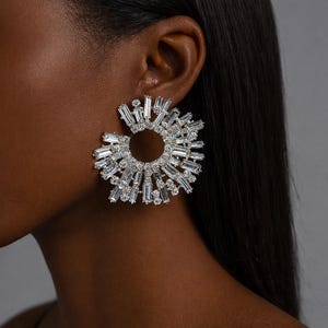 May include: A dazzling, silver-toned earring with a sunburst design. The earring features a circular arrangement of clear, rectangular and round-cut crystals, creating a radiant effect. The jewelry is set against a dark background, highlighting the sparkle of the crystals.