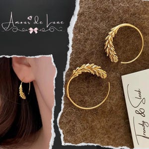 May include: Gold-colored hoop earrings with a wheat stalk design. The earrings are displayed on a brown textured surface. The image also includes the text "Amour de Lune" and "Trendy & Sleek".