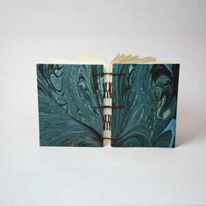 May include: Open journal with a marbled teal and navy cover. The cover design features swirling patterns. The pages are cream-colored and the binding is visible with red thread. The journal is open, displaying the blank pages.