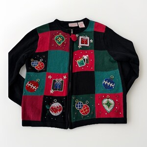 May include: A black zip-up sweater with a patchwork design featuring red, green, and black squares. Each square is decorated with embroidered Christmas ornaments and gift boxes. The sweater has long sleeves and a round neckline.