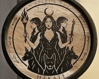 Puede incluir: Posavasos redondo de madera con borde oscuro. El diseño central muestra una mujer con tres caras, una luna creciente y la palabra "HEKATE". El diseño incluye perros, llaves y texto griego.