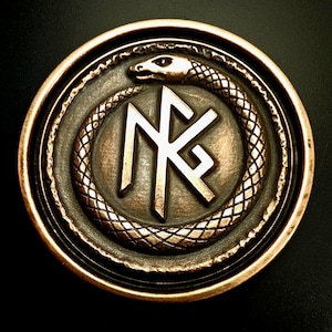 May include: A bronze-colored round emblem featuring a serpent encircling a central design. The serpent's scales are detailed, and the emblem has a raised border. The central design includes stylized letterforms. The overall aesthetic is antique and symbolic.