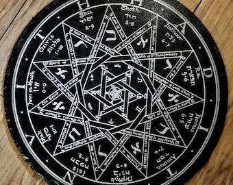 May include: A black, round decorative slate coaster with a white geometric design. The design includes text and symbols arranged in concentric circles and star shapes. The coaster is approximately 8 inches in diameter.