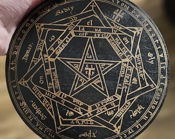 Könnte beinhalten: Eine runde, schwarze Holzplakette mit einem goldenen Pentagramm-Design und umgebendem Text. Die komplizierten Kunstwerke weisen geometrische Formen und Symbole auf und schaffen eine mystische Ästhetik. Die Plakette wird von einer Hand gehalten.