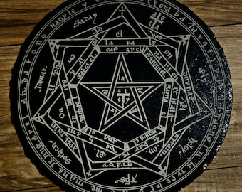 May include: A round, black slate coaster with a white pentagram design and intricate text around the edge. The coaster has a rough, textured surface and sits on a wooden background. The design is reminiscent of occult symbols.