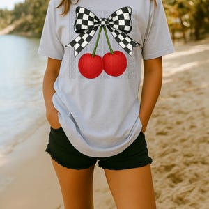 May include: A light gray t-shirt featuring a graphic of two red cherries with green stems and a black and white checkered bow. The shirt is paired with black shorts. The image is taken on a beach.