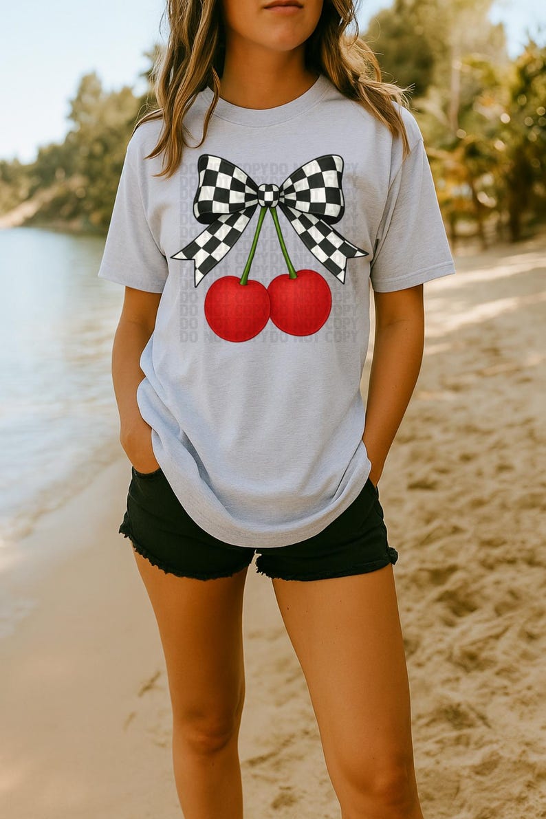 May include: A light gray t-shirt featuring a graphic of two red cherries with green stems and a black and white checkered bow. The shirt is paired with black shorts. The image is taken on a beach.