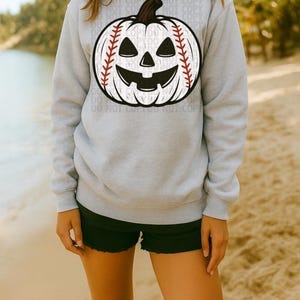 May include: A light gray sweatshirt featuring a baseball-themed jack-o'-lantern design. The pumpkin has a baseball stitch pattern and a carved face. The sweatshirt is a crew neck style.