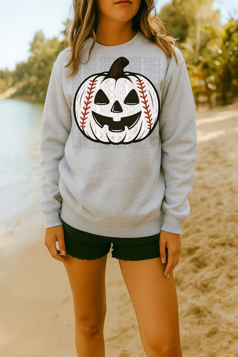 May include: A light gray sweatshirt featuring a baseball-themed jack-o'-lantern design. The pumpkin has a baseball stitch pattern and a carved face. The sweatshirt is a crew neck style.