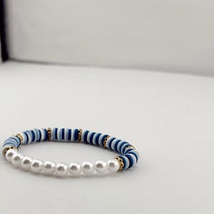 Puede incluir: Una pulsera con cuentas azules, blancas y doradas alternas. La pulsera también incluye una hilera de perlas falsas blancas. La pulsera se muestra sobre un fondo blanco. Las cuentas miden aproximadamente 0,5 cm de diámetro.