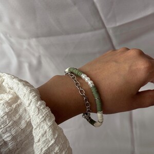Op de afbeelding: Er worden twee armbanden getoond. Eén is een zilveren schakelarmband. De andere armband heeft olijfgroene, witte en parelachtige kralen. De armbanden zitten om de pols van een persoon.