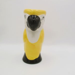 May include: A ceramic tiki mug shaped like a parrot with a glossy finish. The mug features a prominent black beak, a white face, and a bright yellow body. The base of the mug is solid black, providing a stable foundation for the cylindrical vessel.