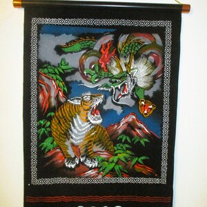 May include: A black fabric wall hanging featuring a vibrant illustration of a roaring tiger and a green dragon. The scene includes red rocky terrain, green foliage, and a dark blue sky. The artwork is framed by a white decorative border pattern.