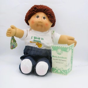 May include: A vintage Cabbage Patch Kid doll with brown yarn hair, wearing a white t-shirt with the text "I Went to Spain" and blue denim pants. The doll holds a birth certificate and a tag.
