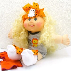 May include: A doll with long curly blonde hair, wearing a grey onesie with a lion graphic and the text "IN WITH THE BIG GUYS". The doll has an orange bow with black polka dots and a ghost patch, and white socks with matching orange bows.