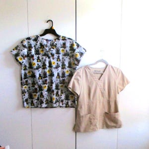 May include: Two short-sleeved medical scrubs hanging on a white background. One scrub is light tan with two front pockets. The other is patterned with black haunted houses, yellow moons, and bats on a light gray background.