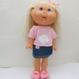 May include: A doll with long blonde hair and a pink bow, wearing a light pink t-shirt with the text CPK and a graphic of a face with the word Smile underneath. The doll is dressed in a dark blue denim skirt and light pink slip-on shoes.