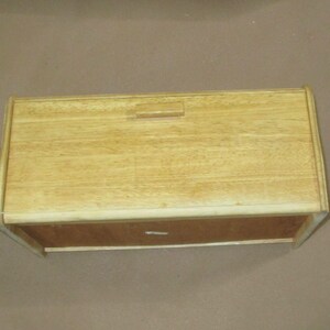 May include: A rectangular wooden bread box with a light tan finish and a visible wood grain pattern. The top features a hinged lid with a small, centered rectangular handle. The front panel is a darker brown wood tone compared to the top lid.