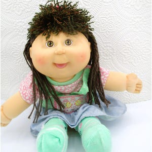 May include: A doll with dark brown curly hair, green eyes, and rosy cheeks. It wears a pink and white patterned short-sleeve shirt under a light blue jumper with a fish graphic, paired with light green socks. The doll has a soft fabric body.