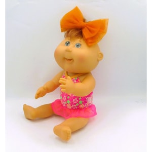 May include: A doll with light skin, round cheeks, and brown hair styled with a large orange sheer fabric bow. The doll is wearing a bright pink swimsuit featuring a colorful floral pattern with green and white accents and a ruffled hemline.