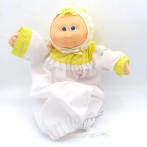 May include: A soft-sculpture doll with a round face, blue eyes, and yarn hair, wearing a white bonnet with yellow gingham trim and lace. The doll is dressed in a white long-sleeved gown featuring a yellow gingham yoke, lace accents, and elastic cuffs.