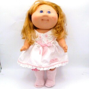 May include: A vintage Cabbage Patch Kids doll with blonde hair and blue eyes. The doll is wearing a white dress with pink accents and a matching bow. It also has pink socks and the words "Baby Real Life" on the dress.