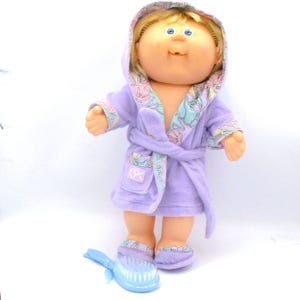 May include: A Cabbage Patch Kids doll wearing a lavender robe with a floral pattern on the trim and matching slippers. The doll has blonde hair and blue eyes. A blue hairbrush is at the doll's feet.
