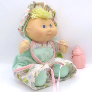 May include: A doll with a soft vinyl head, yellow yarn hair, and blue eyes. It wears a mint green and white striped outfit with a matching bonnet featuring a small floral print. A small pink plastic baby bottle is attached by a thin pink ribbon.