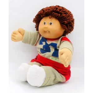 May include: A vintage Cabbage Patch Kid doll with brown yarn hair, a tan face, and blue eyes. The doll wears a tan and red outfit with the number 27 in blue on the front. The doll is sitting with its arms outstretched.