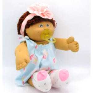 May include: A soft-bodied doll with dark brown yarn hair, wearing a light pink headband with a large fabric flower. The doll has a yellow pacifier in its mouth and wears a light blue sleeveless dress with a pink and white floral pattern.