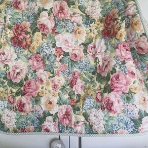 May include: A quilted fabric featuring a dense floral pattern with pink, red, and yellow roses interspersed with small blue flowers and green leaves on a pale yellow background, finished with a thin green and white checkered fabric border.