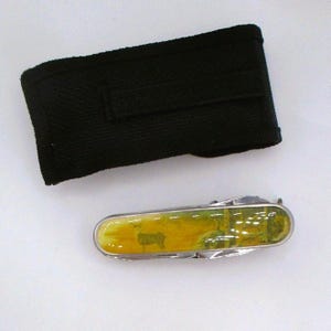 May include: A silver multi-tool with a yellow and green landscape design featuring a deer. The multi-tool is next to a black nylon carrying case. The multi-tool has several folding blades and tools.
